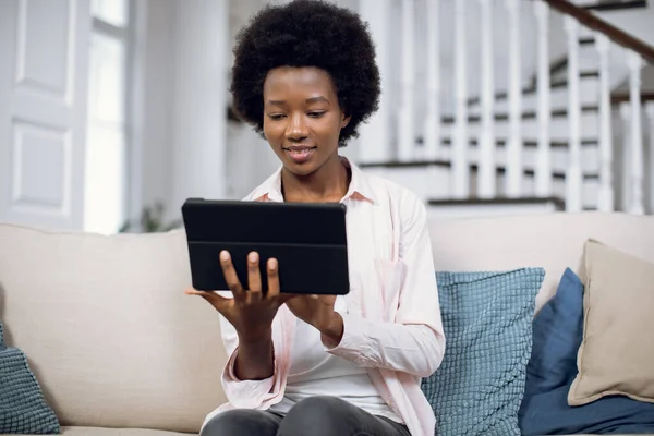 Sorridente bella giovane donna africana utilizzando tablet sul comodo divano in soggiorno — Foto Stock