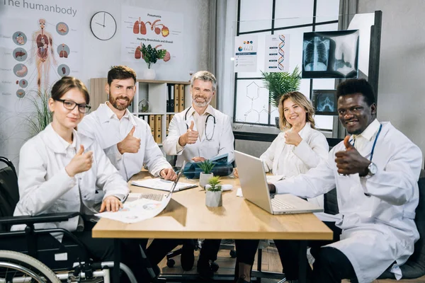 Пять практикующих врачей разных национальностей встречаются в офисе и показывают большие пальцы — стоковое фото