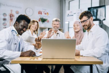 Tekerlekli sandalyedeki nitelikli doktorlar ofiste video görüşmesi için dizüstü bilgisayar kullanıyorlar. Çevrimiçi konferans sırasında gülümseyen ve el sallayan bir grup çok ırklı tıp uzmanı.