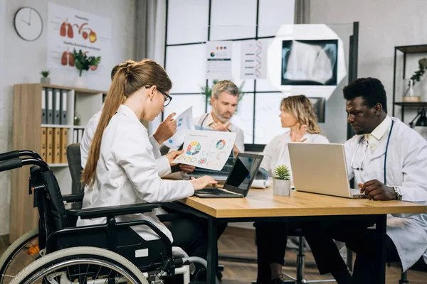 Tekerlekli sandalyedeki kadın doktor çok ırklı meslektaşlarıyla konferans sırasında kablosuz bilgisayarla çalışıyor. Ortak tıbbi araştırmalarda ilerleme sağlamak için işbirliği yapan bir grup terapist..