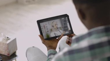 Afrikalı erkek hasta, elinde tabletle evde oturuyor, GPS doktoruyla görüntülü konuşma yapıyor.