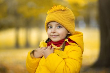 Sarı ceketli, kırmızı eşarplı, güz ağaçlarının arka planında kameraya gülümseyerek poz veren sevimli bir kızın açık hava portresi. Sarı yapraklar kızların kafasına ve omuzlarına düştü..
