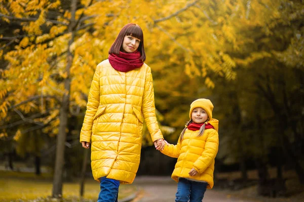 Mother and daughter walking in autumn yellow park. Happy mom and little girl in yellow jackets holding hands and enjoying walk outdoors in the autumn.