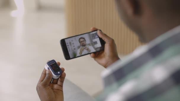 Hombre africano se sienta con el teléfono celular y sostiene el pulso del dedo oxímetro, tener chat de vídeo con el médico masculino — Vídeos de Stock