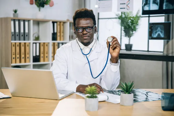 Medico africano seduto in ufficio e che tiene lo stetoscopio — Foto Stock