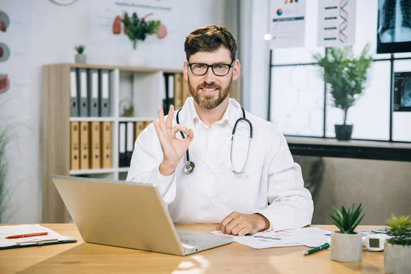 Bello maschio medico mostrando ok segno e sorridente sulla macchina fotografica in ufficio — Foto Stock