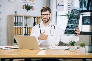 Masasında oturan erkek doktor ellerinde röntgen taramasıyla baş parmağını gösteriyor.