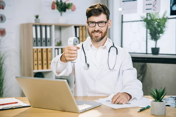 Симпатичный семейный врач сидит за столом с современным электронным термометром в руках — стоковое фото