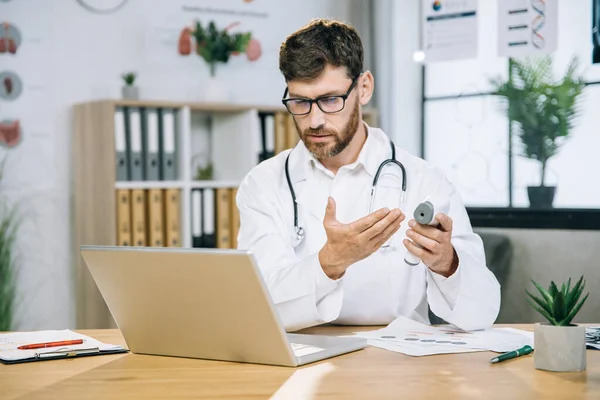 Kaukázusi orvos nézi a laptopot és tartja a hőmérőt — Stock Fotó