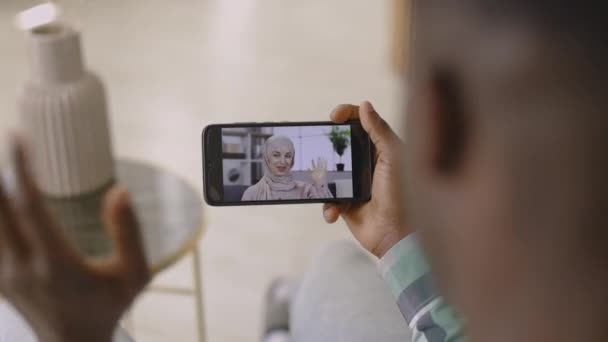 Homme africain, assis sur le canapé avec téléphone, agitant la main, ayant un appel vidéo avec un ami musulman — Video