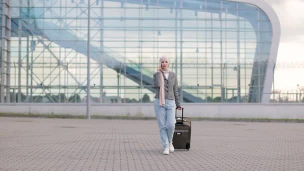 Muslim woman in hijab, walking outdoors carrying a suitcase and talking phone — Stock Video