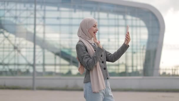 Mujer musulmana con pañuelo en la cabeza, haciendo videollamadas con un amigo hablando y señalando el edificio del aeropuerto — Vídeo de stock