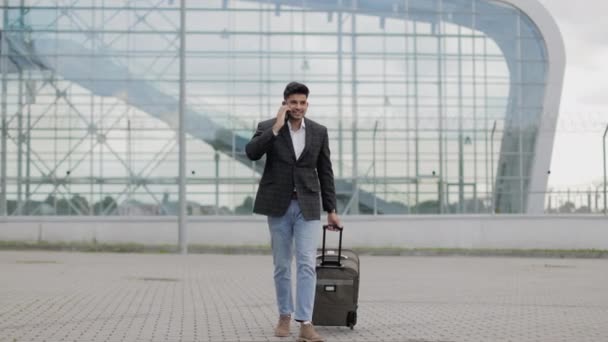 Арабский бизнесмен разговаривает по телефону, гуляя за пределами современного аэропорта, с чемоданом — стоковое видео