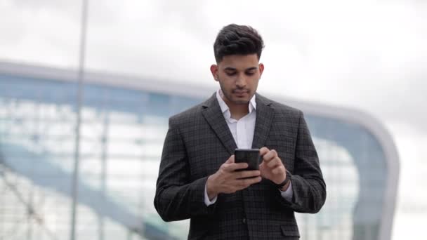 Empresário indiano em jaqueta formal, de pé perto do edifício moderno e mensagens de texto no smartphone — Vídeo de Stock