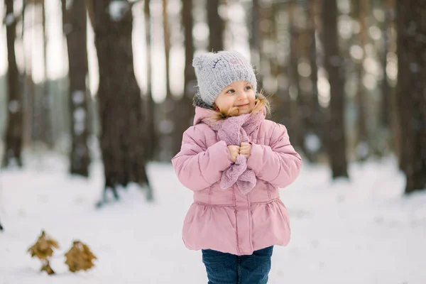 Winter in the forest. Cute dreamy little girl in a pink coat standing in snowy forest with trees background and looking away. Smiling kid walking in frosty winter day in forest — Stock Photo, Image