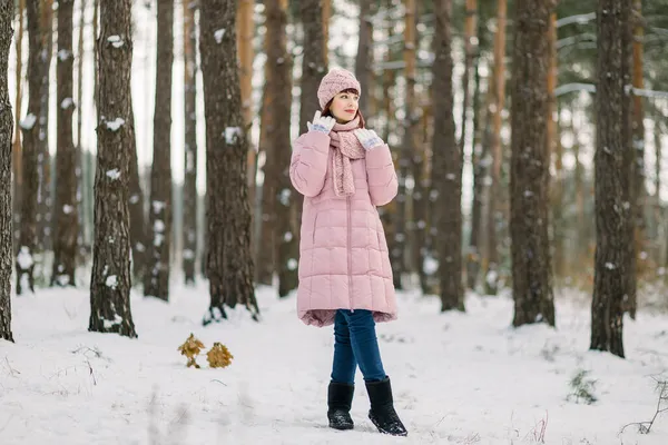 Sonhador encantador jovem caucasiana em roupa rosa elegante, de pé sobre o fundo de pinheiros na floresta de inverno nevado e olhando para longe com sorriso. — Fotografia de Stock