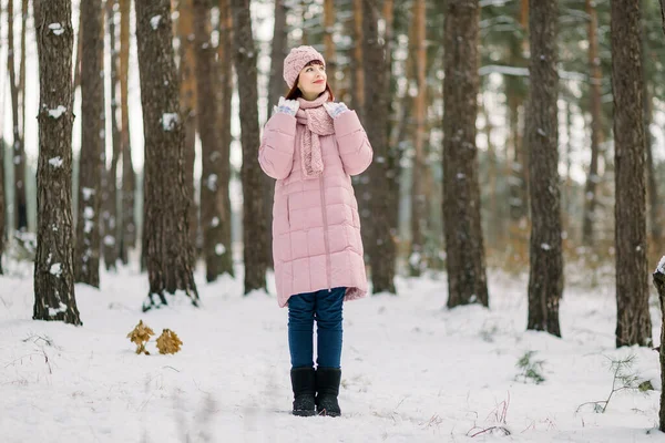 Sonhador encantador jovem caucasiana em roupa rosa elegante, de pé sobre o fundo de pinheiros na floresta de inverno nevado e olhando para longe com sorriso. — Fotografia de Stock