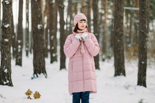 Outdoor winter portrait of pretty young woman wearing pink coat and stylish knitted hat and scarf, standing in beautiful snowy forest in front of pine trees and enjoying walk on frosty day. — Stock Photo, Image