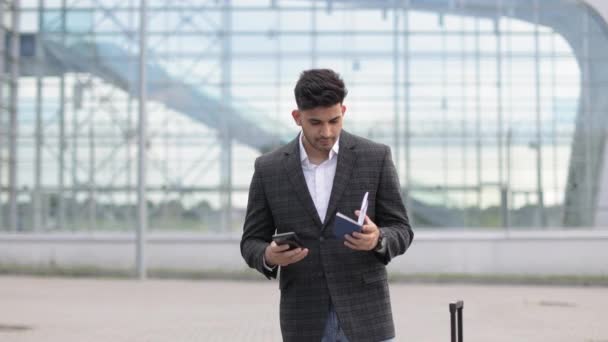 Sympathischer junger Inder mit Mobiltelefon, der vor dem Flughafenterminal steht und seinen Reisepass hält — Stockvideo