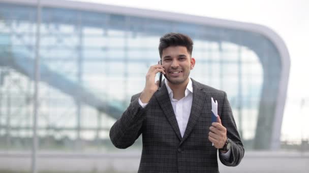 Arabski biznesmen, rozmawiający przez telefon, trzymający paszport i bilet, stojący przed lotniskiem — Wideo stockowe
