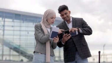 Arap çift, havaalanı terminalinin önünde bavullarıyla duruyor ve uçuş kartlarını kontrol ediyorlar.