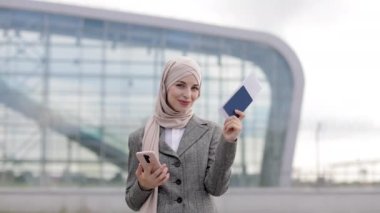 Havaalanı terminalinin önünde duran tesettürlü Müslüman kadın telefon ve pasaport tutuyor.