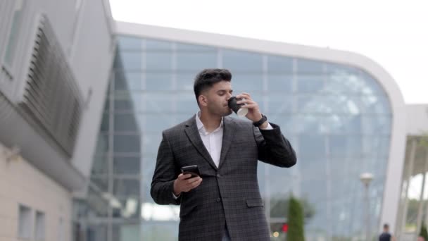 Індійський бізнесмен стоїть перед сучасним будинком, розмовляє по мобільному телефону, п "є каву. — стокове відео