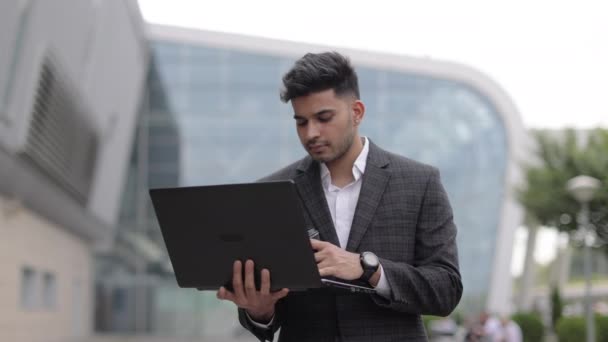 Улыбающийся индус позирует на камеру во время работы над ноутбуком на открытом воздухе офиса — стоковое видео