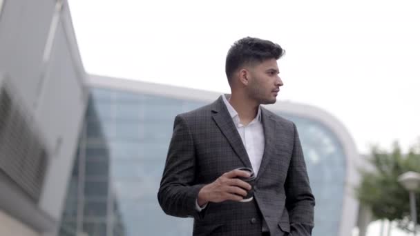Молодой вдумчивый араб, стоящий у современного здания и пьющий, забирает кофе — стоковое видео