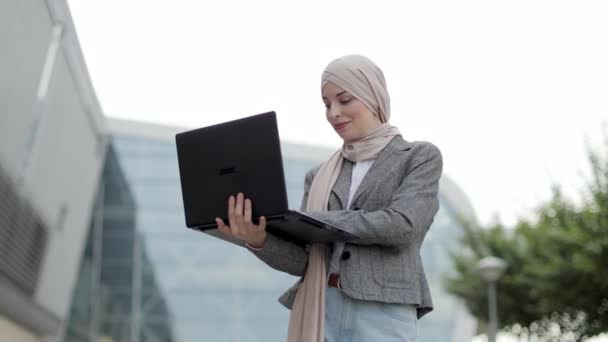 Glimlachende moslim dame in hijab poseren op camera tijdens het werken op laptop buiten kantoor — Stockvideo