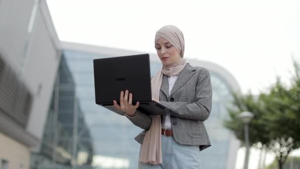 Mulher muçulmana em desgaste casual inteligente, de pé fora do edifício moderno e internet surf no laptop — Vídeo de Stock