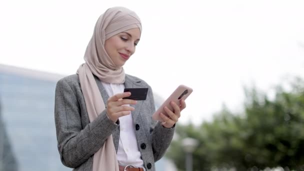 Мусульманка в хиджабе, держит смартфон и кредитку, делает покупки на улице — стоковое видео
