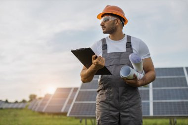Üniformalı mühendis güneş panellerinin arasında dikiliyor ve başka tarafa bakıyor.
