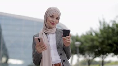 Başörtülü Arap kadın kahve ve telefonla sokakta yürüyor.