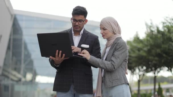 Het concept mens en technologie. Aantrekkelijk zakenkoppel van Arabische man en moslimvrouw in slimme casual stijl, buiten kantoor staan, samenwerken op laptop, lachen en discussiëren. — Stockvideo