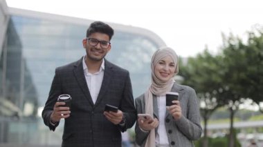 Gülümseyen iş çifti, yakışıklı Arap erkek ve tesettürlü Müslüman kadın, kahve molası için sokakta duruyorlar, telefon kullanıyorlar ve kahve içiyorlar..