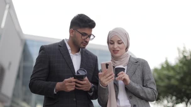 Árabe pareja de negocios divirtiéndose y hablando juntos caminando por la calle durante la pausa para el café. Mujer en hijab mostrando la pantalla de su smartphone a su colega, compartiendo ideas o noticias. — Vídeos de Stock