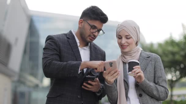 Zakenmensen bij koffiepauze buiten. Gelukkige sympathieke hindoe man praten met zijn collega kantoor, mooie moslim vrouw in hijab en iets laten zien op de smartphone. Koffie drinken samen. — Stockvideo