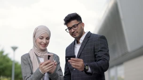 Conceptual photo of hindu businessman showing credit card number to his female Muslim colleague typing on smartphone and making online payment. Happy people outdoors shopping online. — Stock Video