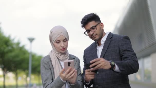 Zwei multiethnische Geschäftsleute, die im Freien Finanztransaktionen durchführen. Muslimische Dame im Hidschab nutzt Smartphone für bargeldloses Bezahlen, ihr arabischer männlicher Kollege hält Kreditkarte. — Stockvideo