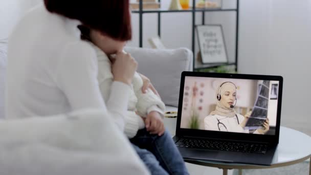 Vista posterior de mamá midiendo la temperatura de una niña enferma y teniendo video chat con la mujer árabe médico en el ordenador portátil — Vídeo de stock