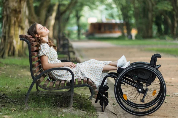 Relaxed woman sitting on bench with legs on wheelchair — Stockfoto