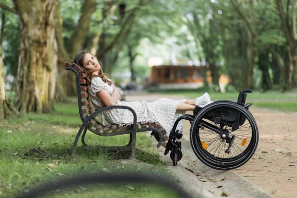 Γυναίκα με ειδικές ανάγκες που κάθεται στον πάγκο κοντά στην αναπηρική καρέκλα της στο πάρκο — Φωτογραφία Αρχείου