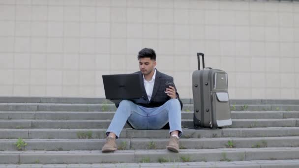Młody arabski biznesmen pracujący na laptopie, siedząc na schodach na ulicy, z walizką, czekając na podróż służbową i pijąc kawę. — Wideo stockowe