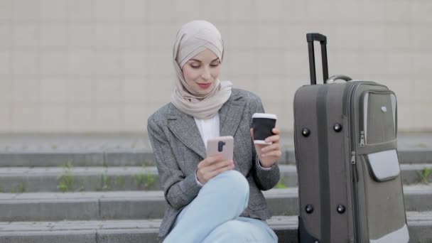Giovane donna musulmana in hijab vestita con jeans e giacca utilizzando il telefono e bevendo caffè take away, mentre seduto sulle scale con borsa da viaggio. — Video Stock