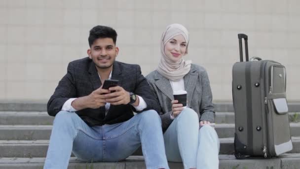 Cheerful smiling diverse business man and woman in hijab sitting on the stairs outdoors with smarphones and coffee, waiting for their business trip. Big travel bag on the stairs. — Stockvideo