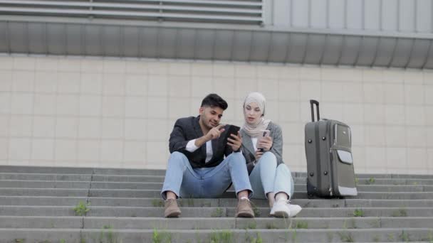 Angenehm lächelnder indischer Mann und hübsche muslimische Dame im Hijab, die mit Reisekoffer auf der Treppe sitzen und ihre Abfahrtszeit überprüfen. Geschäftspaar sitzt auf der Treppe vor dem Flughafen. — Stockvideo