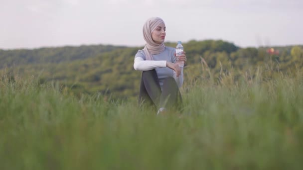 Happy smiling muslim woman in hijab with water bottle sitting on mat on beautiful park at morning — Stock Video