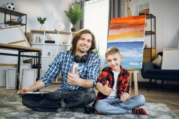 Son with father showing thumbs up while sitting near easel — Foto de Stock