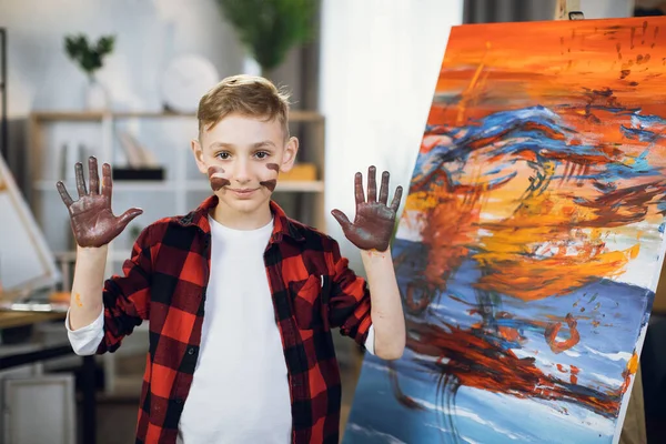 Niño con pintura en las mejillas y las manos posando cerca del caballete —  Fotos de Stock